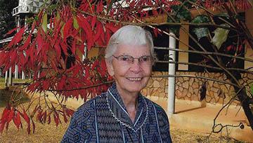 Sr. Kathleen Feeley wears a Navy blue print shirt surrounded by red leaves from a tree