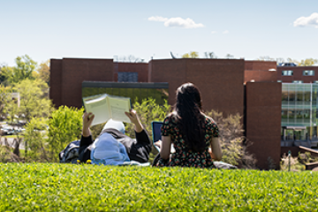 2 students sitting in the grass reading with the library in the distance