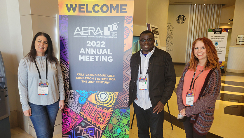 Three NDMU students and faculty pose next to a welcome side at the AERA conference.