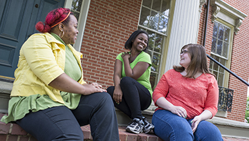 Three women students sit together outside in front of a brick building