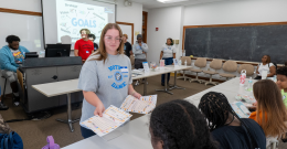 Female student wearing light blue NDMU t-shirt stands in front of a table with children facing her