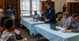 Male student stands at a table with other seated male students in front of seated children