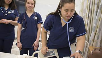 Three female students wearing Navy blue nursing scrubs in a hospital-like setting, and one student uses a stethoscope on a mannikin