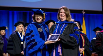 Mary Packard wearing regalia receives award plaque from President Yam
