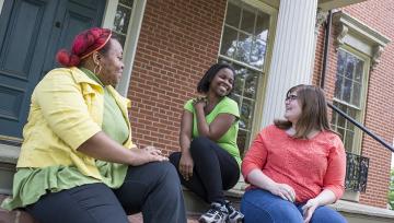 Three women students sit together outside in front of a brick building