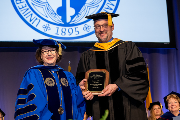 James Culhane wearing regalia holds a plaque as he stands with President Yam