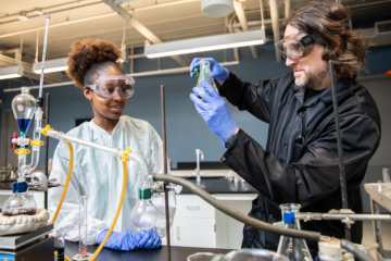 Female student wearing a white lab coat in a lab with a male professor