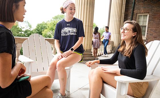 Three female students smile and engage with each other while sitting on white Adirondack chairs on a balcony