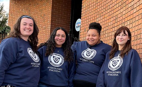 Four female students wearing Navy blue sweatshirts pose together in front of a brick wall