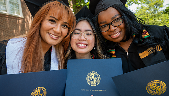 three students in caps and gowns smiling holding diplomas