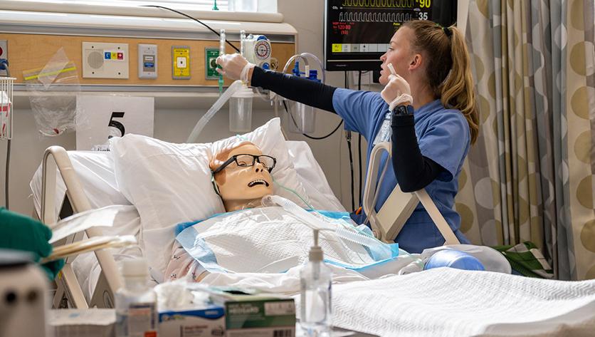 Female student wearing light blue scrubs in a nursing simulation room with a manikin