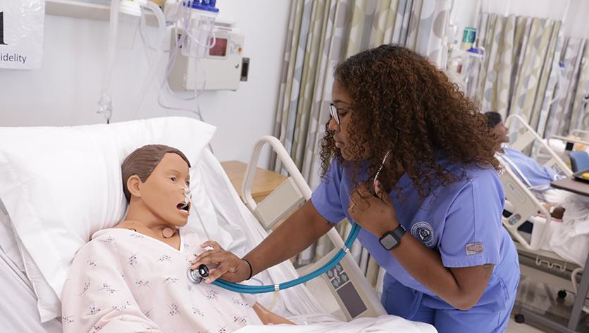 Female wearing blue nursing scrubs checks temperature on mannikin in a nursing simulation lab