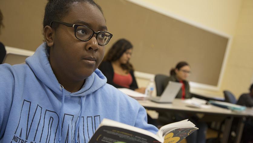 Female student wearing light blue NDMU hoodie holds a book in a classroom