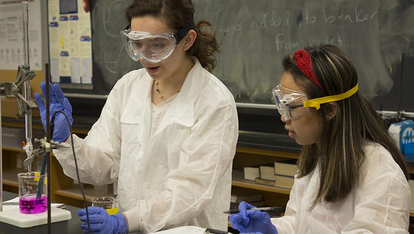 Two female students wearing white lab coats work together in a lab