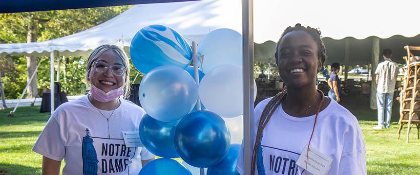 Two female students wearing white t-shirts smile as they stand next to blue and white balloons