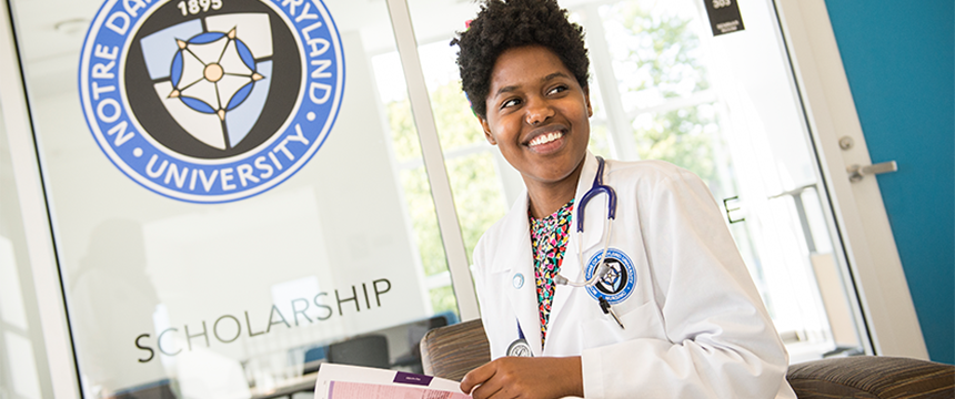 African American female nurse wearing NDMU lab coat in front of the University logo