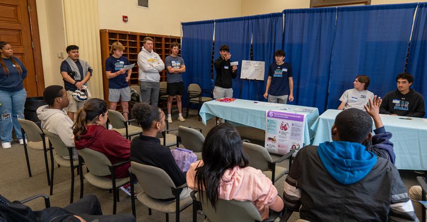 A group of male students wearing NDMU gear stand or sit in front of a blue curtain and speak to a group of seated children