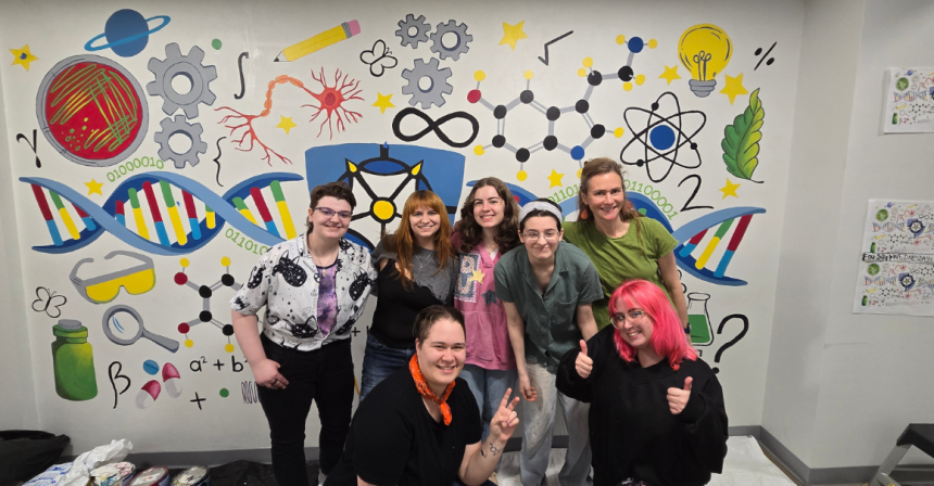 Female students pose while standing in front of the STEAM mural