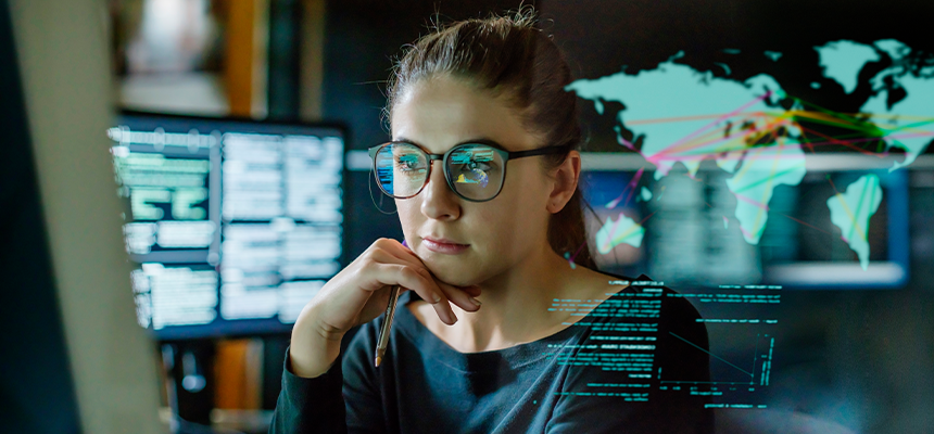 young woman, wearing glasses, surrounded by computer monitors in a dark office. In front of her there is a see-through displaying showing a map of the world with some data.